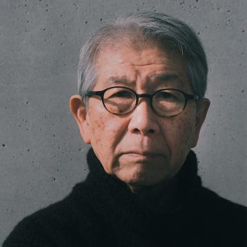 Foto do arquiteto Riken Yamamoto com óculos e blusa de gola longa preta na frente de parede cinza