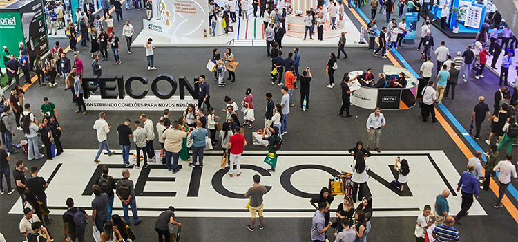 Foto aérea da entrada da exposição Feicon 2024. No chão, o nome da feira recobre todo o piso. Há muitas pessoas, espalhadas pela área central. Na parte superior da foto, começam a aparecer os estandes.
