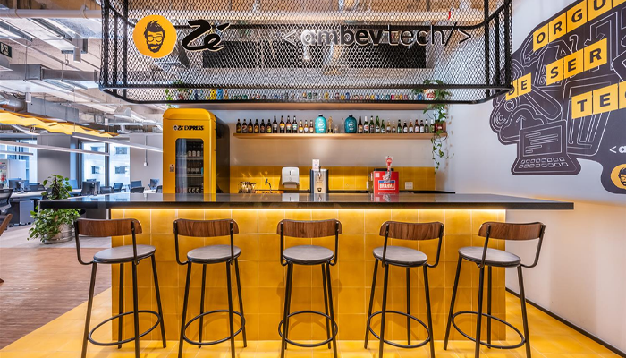 Foto da área de recepção da empresa do Zé Delivery/Ambev Tech, que simula um bar. A meia parede foi revestida por azulejos amarelos, há banquetas em frente ao balcão e, no topo, o logo do aplicativo 