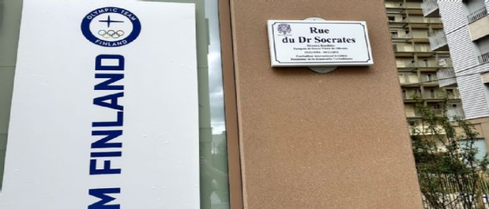 Placa Rue du Dr Socrates inserida em parede marrom de prédio escrito Finland em azul escuro, contendo ao fundo prédios e galhos.