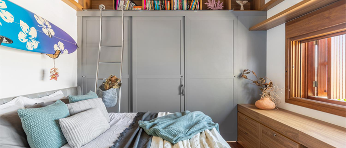 Quarto com cama, guarda-roupa, cômoda e bancada de estudos em frente da cama. Destaque para prancha de surfe pendurada na parede